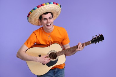 Young man in Mexican sombrero hat playing guitar on violet background