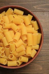 Raw rigatoni pasta in bowl on wooden table, top view