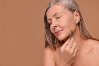 Photo of Woman massaging her face with jade gua sha tool on brown background, space for text