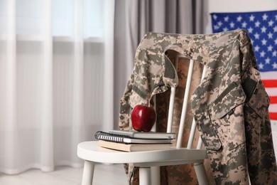 Photo of Chair with soldier uniform, notebooks and apple near flag of United States indoors, space for text. Military education