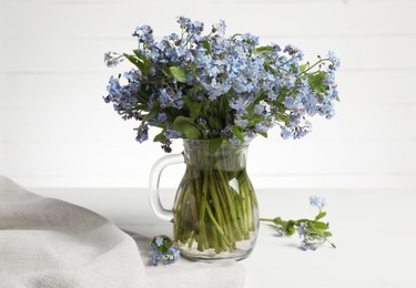 Bouquet of beautiful forget-me-not flowers in glass jug and blue cloth on white table