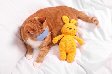 Photo of Cute ginger cat with sleep mask and crocheted bunny resting on bed