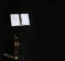 Trumpet, case and note stand with music sheets on black background. Space for text