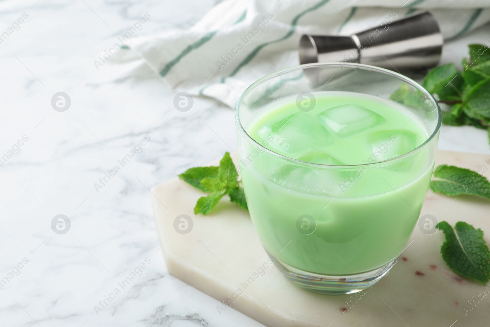 Photo of Delicious mint liqueur with ice cubes and green leaves on white marble table, closeup. Space for text