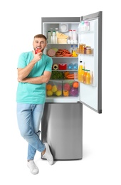 Photo of Young man eating apple near open refrigerator on white background