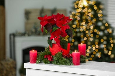 Potted poinsettia, burning candles and festive decor on white dresser in room. Christmas traditional flower