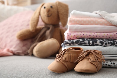 Toy bunny, stack of stylish child clothes and shoes on sofa