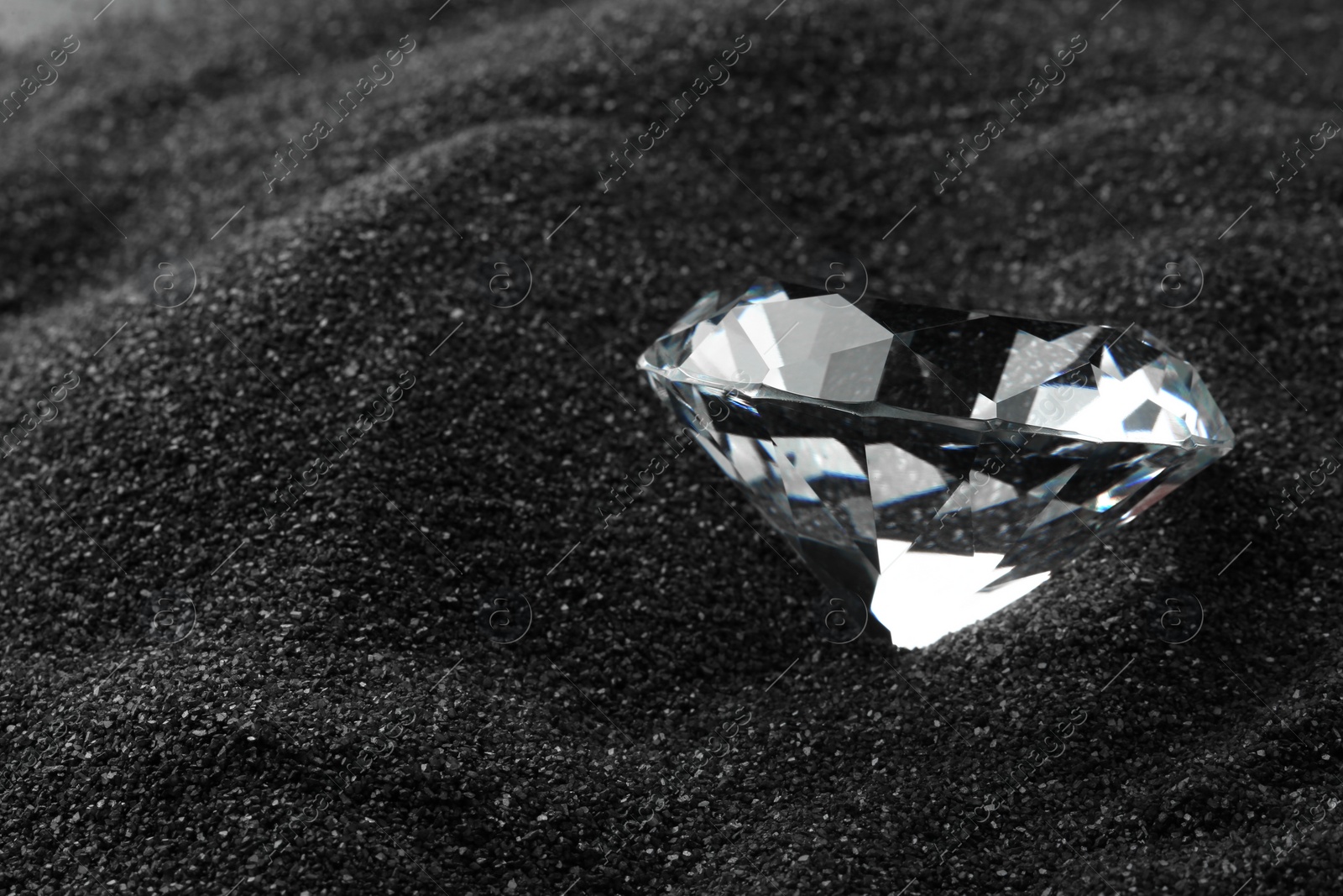 Photo of Beautiful shiny diamond on decorative black sand. Space for text