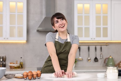 Happy young housewife kneading dough at white table in kitchen