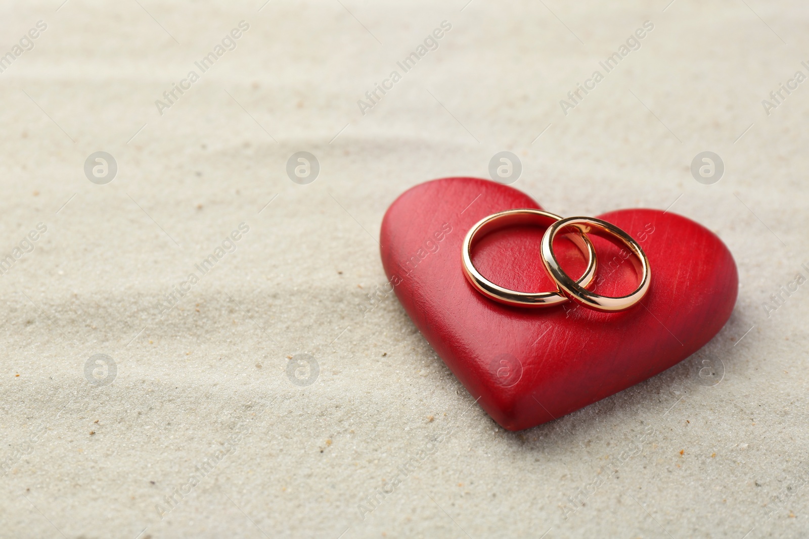 Photo of Honeymoon concept. Two golden rings and red wooden heart on sand