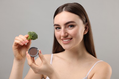 Photo of Smiling woman making fake freckles with broccoli and cosmetic product on grey background