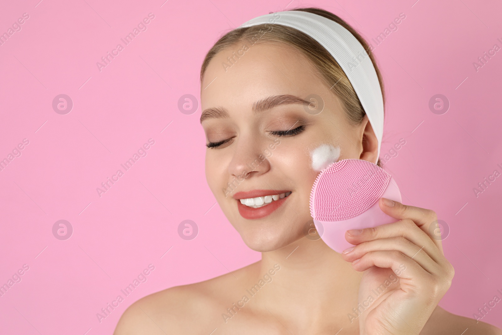 Photo of Young woman washing face with brush and cleansing foam on pink background. Cosmetic products