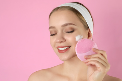 Photo of Young woman washing face with brush and cleansing foam on pink background. Cosmetic products