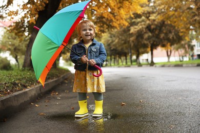 Photo of Cute little girl with colorful umbrella standing in puddle outdoors