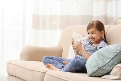 Cute little girl with glass of milk on sofa at home
