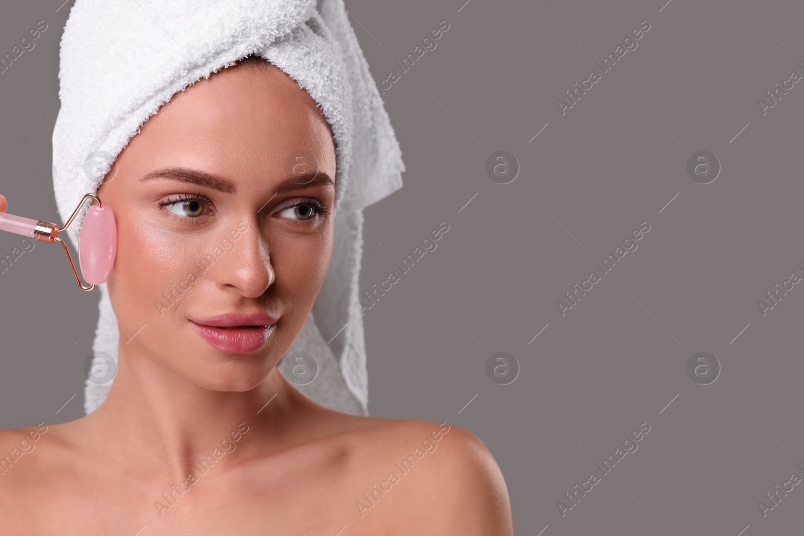 Photo of Young woman massaging her face with rose quartz roller on grey background, space for text