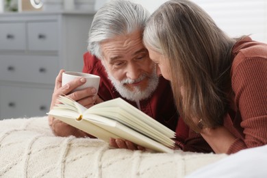 Photo of Senior man with cup of drink and his wife reading book on bed at home
