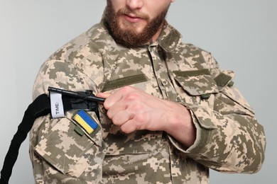 Photo of Ukrainian soldier in military uniform applying medical tourniquet on arm against light grey background, closeup