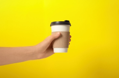 Woman holding takeaway paper coffee cup with cardboard sleeve on yellow background, closeup