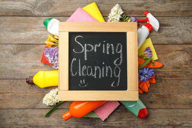 Photo of Blackboard with words Spring Cleaning, flowers and detergents on wooden table, flat lay