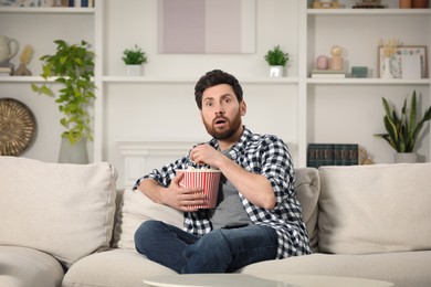 Photo of Surprised man watching TV with popcorn on sofa at home