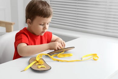 Little boy tying shoe lace using training cardboard template at white table indoors, space for text