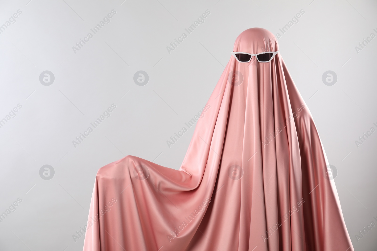 Photo of Glamorous ghost. Woman in pink sheet with sunglasses on light grey background, space for text