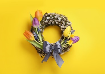 Willow wreath with different beautiful flowers and grey bow on yellow background, top view