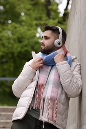 Photo of Handsome man in warm scarf and headphones near wall outdoors