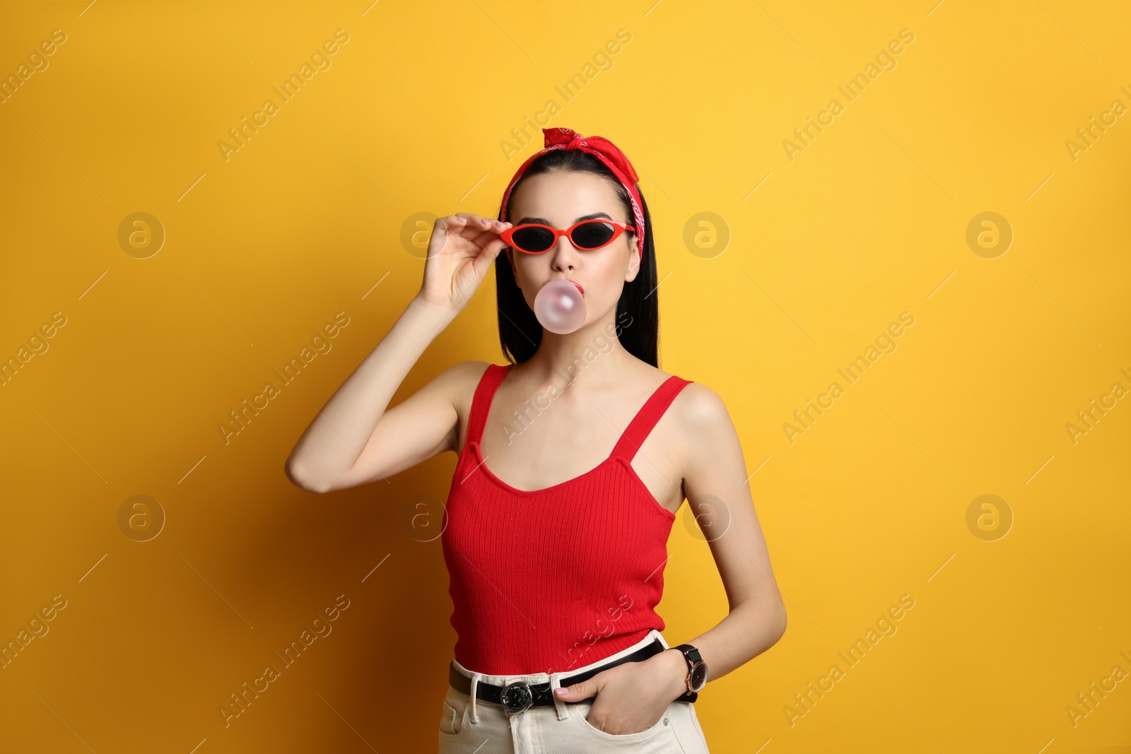 Photo of Fashionable young woman in pin up outfit blowing bubblegum on yellow background