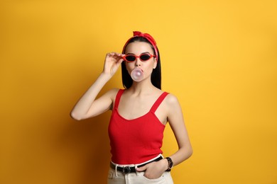 Fashionable young woman in pin up outfit blowing bubblegum on yellow background