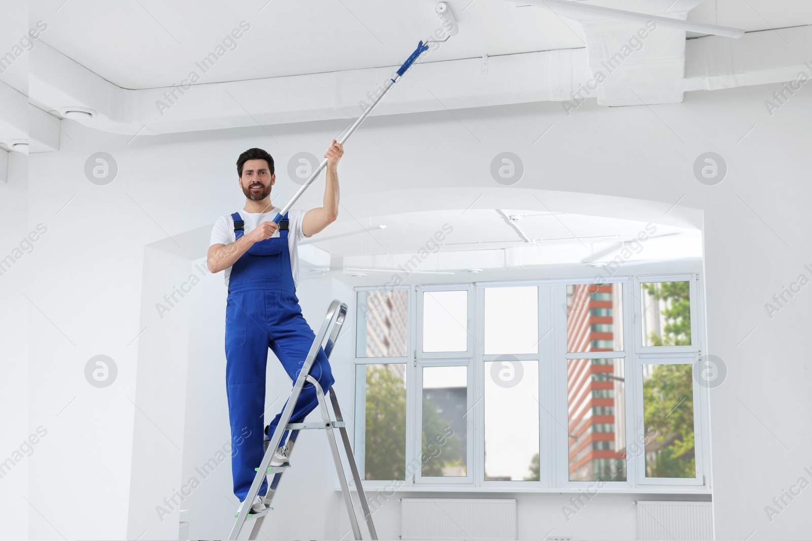 Photo of Handyman painting ceiling with roller on step ladder in room