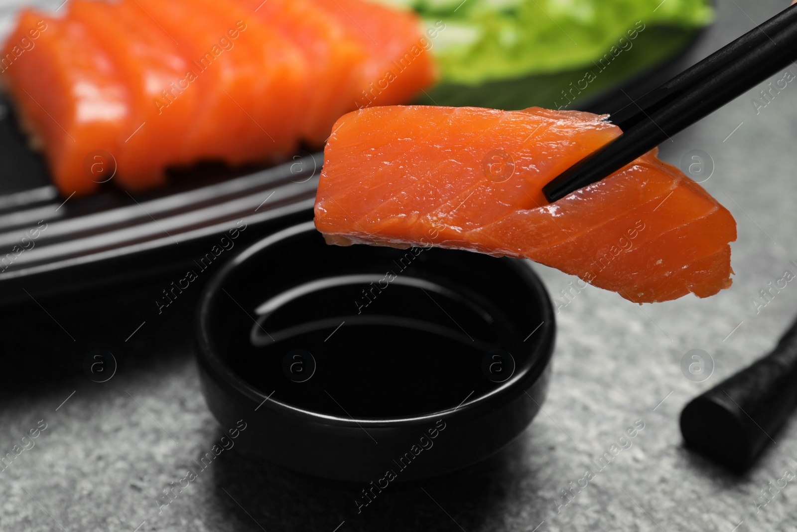 Photo of Dipping tasty salmon slice into soy sauce with chopsticks at grey table, closeup. Delicious sashimi dish