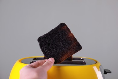 Woman holding burnt bread near toaster against grey background, closeup