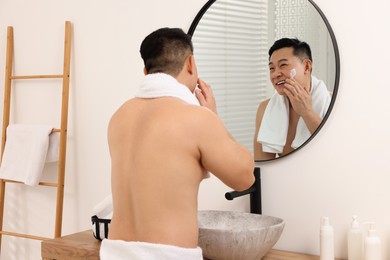 Photo of Handsome man applying cream onto his face near mirror in bathroom