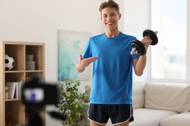 Photo of Smiling sports blogger holding dumbbell while recording fitness lesson with camera at home