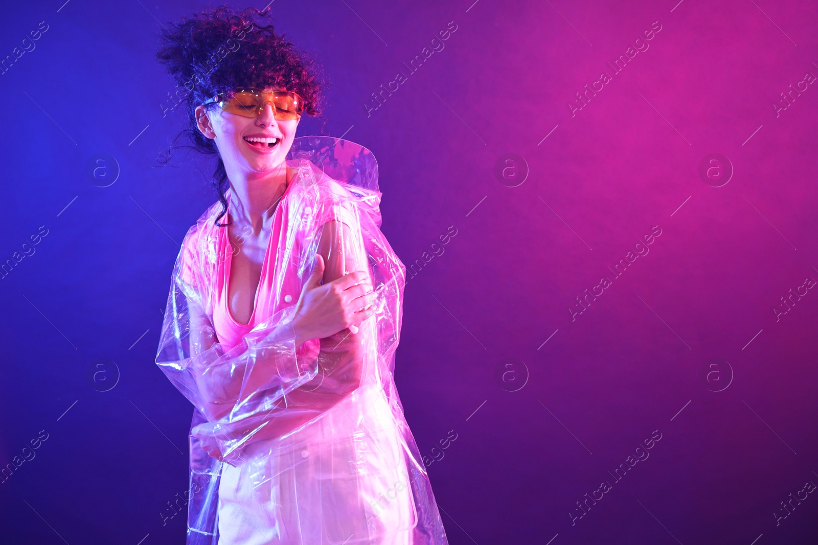 Photo of Beautiful young woman in transparent coat and sunglasses posing on color background in neon lights. Space for text