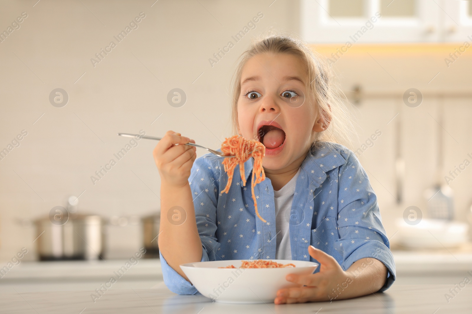 Photo of Cute little girl eating tasty pasta at table in kitchen