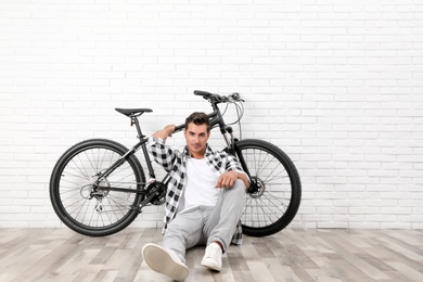 Handsome young man with modern bicycle near white brick wall indoors