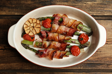 Photo of Oven baked asparagus wrapped with bacon in ceramic dish on wooden table, top view