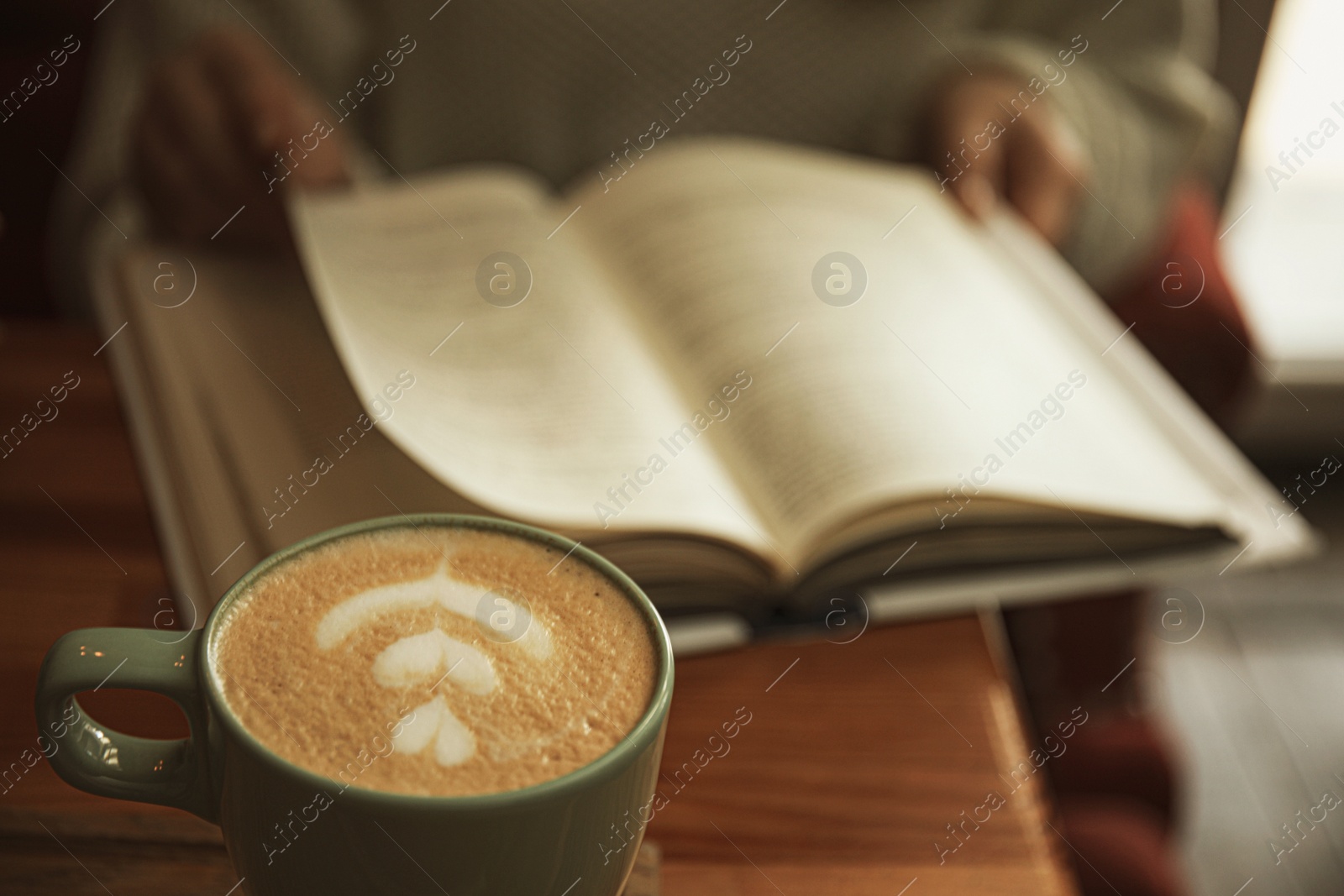 Photo of Woman with coffee reading book indoors, focus on cup