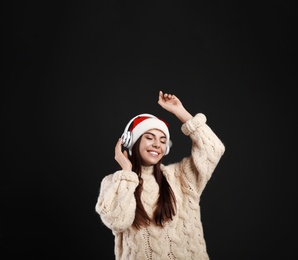 Young woman in Santa hat listening to Christmas music on black background