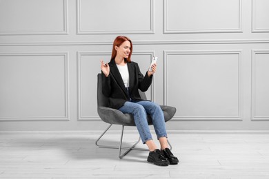 Happy young woman having video chat via smartphone while sitting in armchair near light grey wall indoors