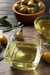 Photo of Glass bowls of oil, ripe olives and green leaves on wooden table