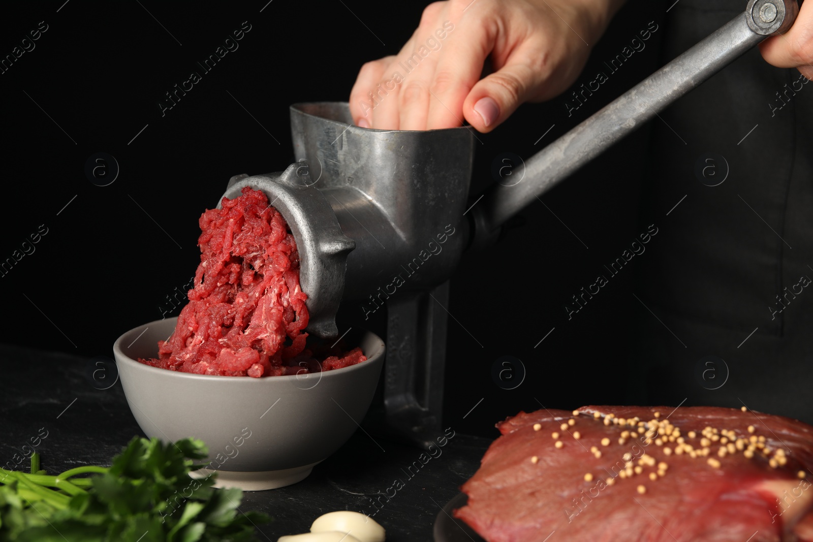 Photo of Woman making beef mince with manual meat grinder at table against black background, closeup