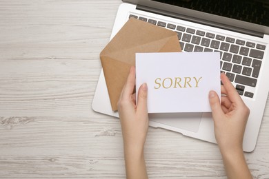 Image of Apology. Woman holding card with word Sorry near envelope and laptop at white wooden table, top view. Space for text