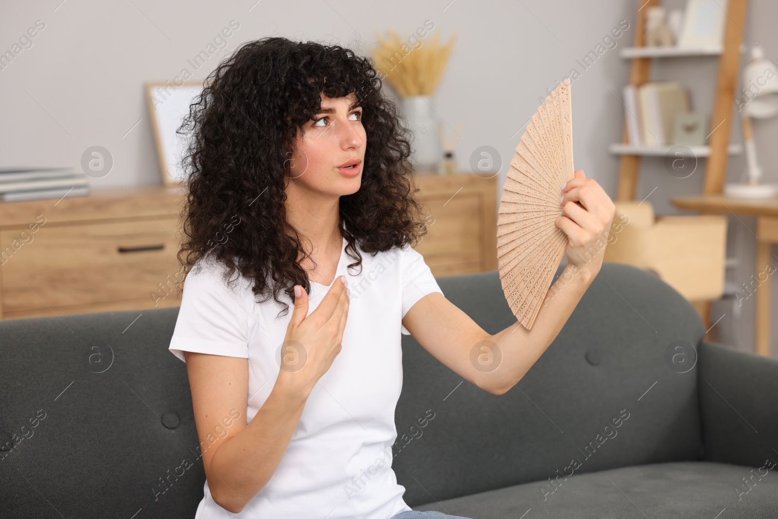 Photo of Young woman waving hand fan to cool herself on sofa at home