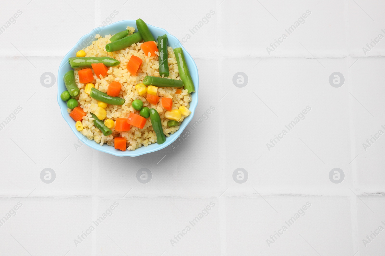 Photo of Delicious bulgur with vegetables in bowl on white tiled table, top view. Space for text