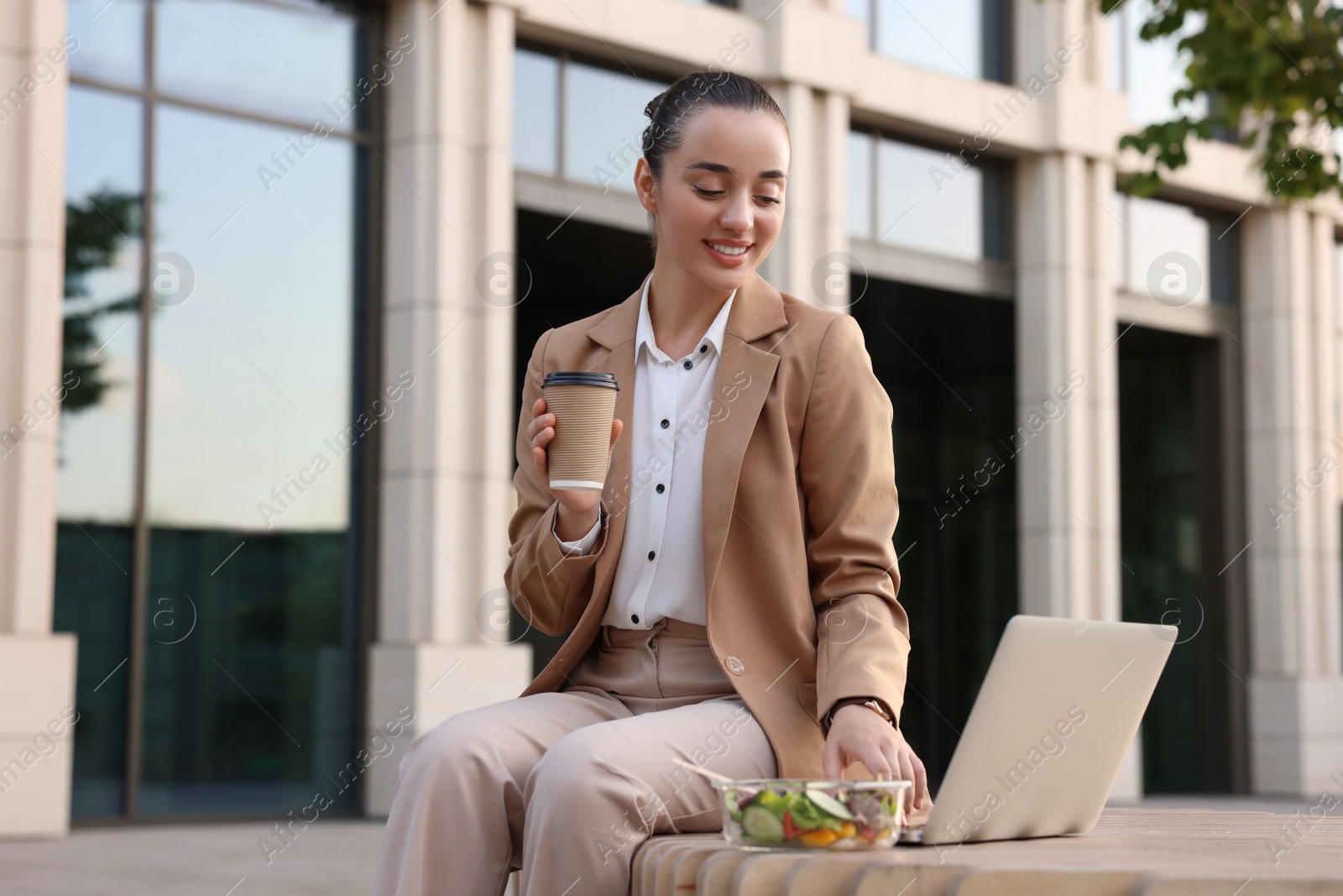 Photo of Happy businesswoman with paper cup of coffee using laptop during lunch on bench outdoors