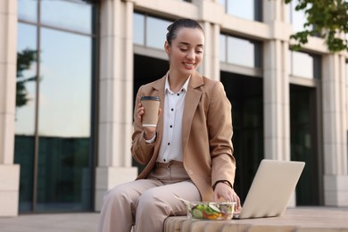 Photo of Happy businesswoman with paper cupcoffee using laptop during lunch on bench outdoors